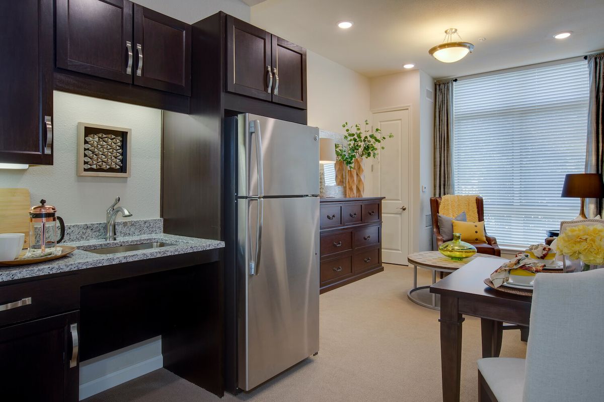 A modern apartment kitchen and living area with dark cabinets, a stainless steel refrigerator, and contemporary furnishings.