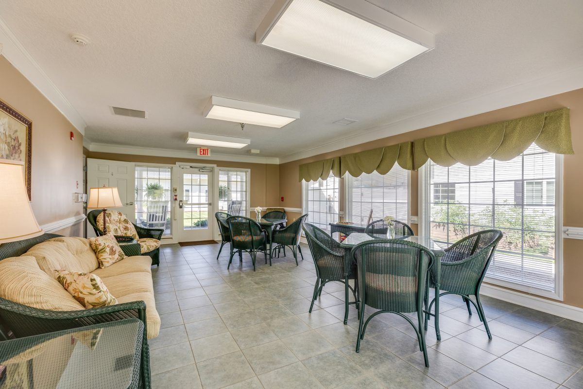 A well-lit community room featuring tables and chairs, with large windows allowing natural light.