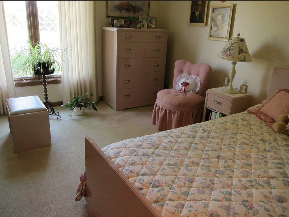 A cozy bedroom featuring a patterned bedspread, pink chair, and large dresser near a window with blinds.