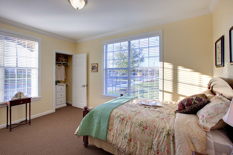 A cozy bedroom with natural light streaming through a large window, featuring a neatly made bed and soft carpeting.