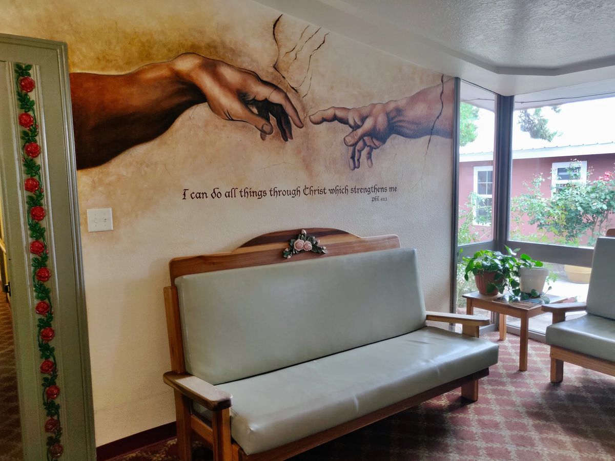 A mural featuring two hands reaching towards each other, inspired by a classic artwork, adorns the wall above a wooden bench in a cozy indoor space with a window and potted plant.