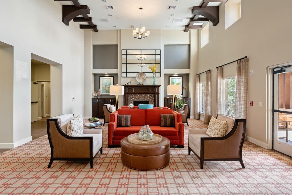 A spacious living room with a red sofa and armchairs, centered around a circular coffee table. A chandelier hangs from the ceiling, and natural light pours in through large windows.