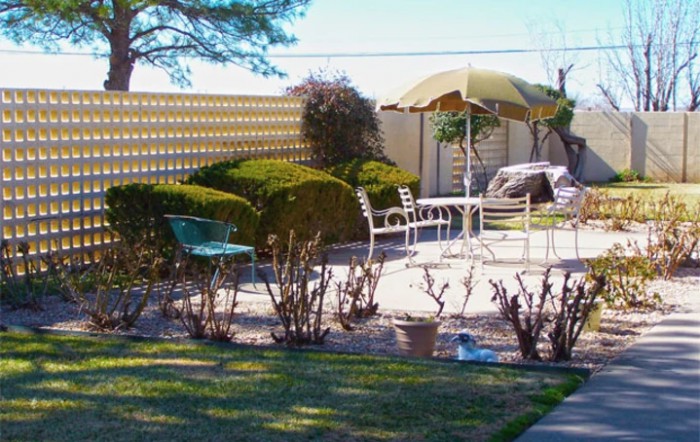 A sunlit patio area with outdoor furniture and an umbrella, surrounded by neatly trimmed bushes and a garden.