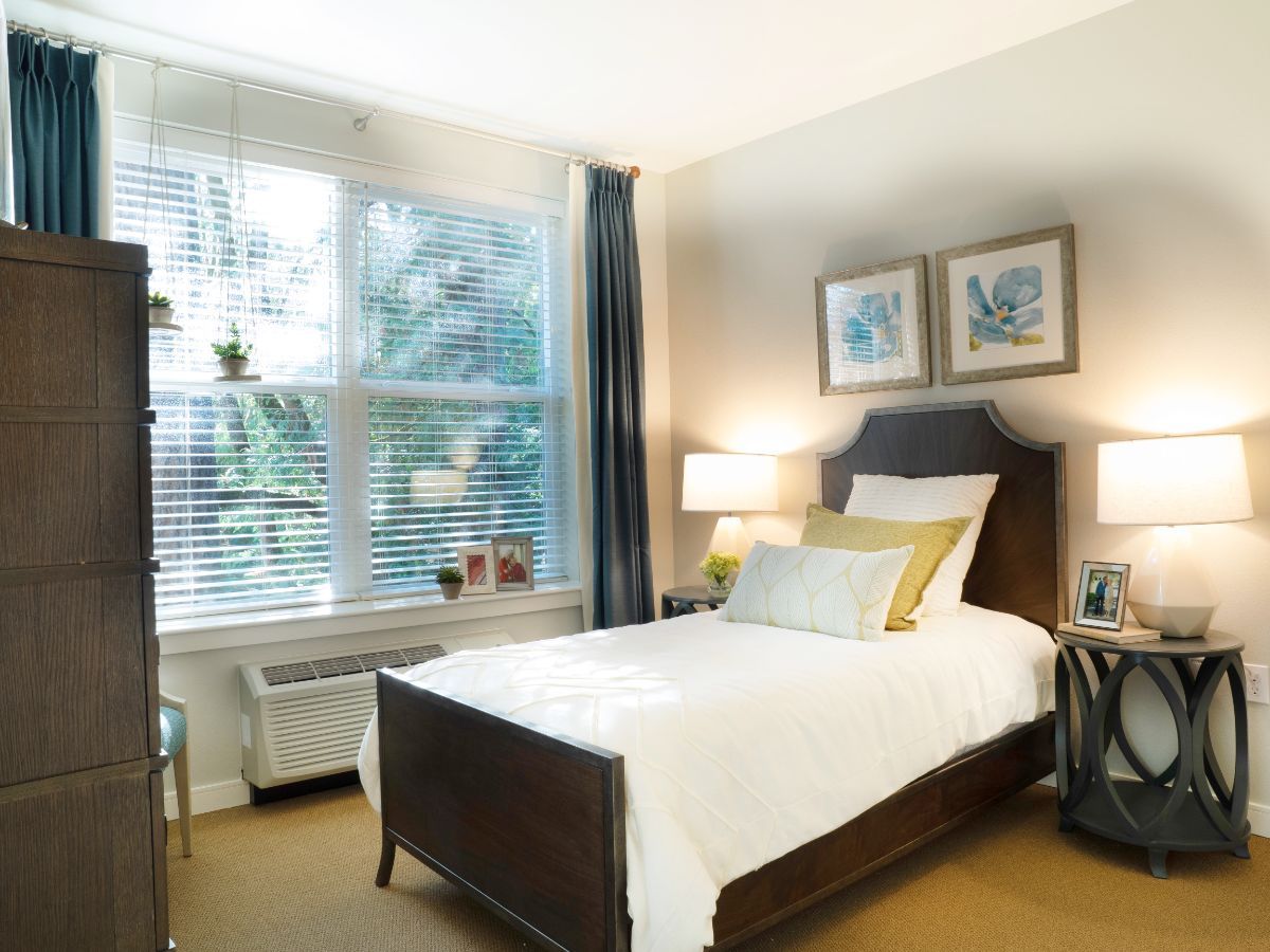 A cozy bedroom with a single bed, decorated with white bedding and accent pillows, featuring artwork on the wall and a large window allowing natural light in.