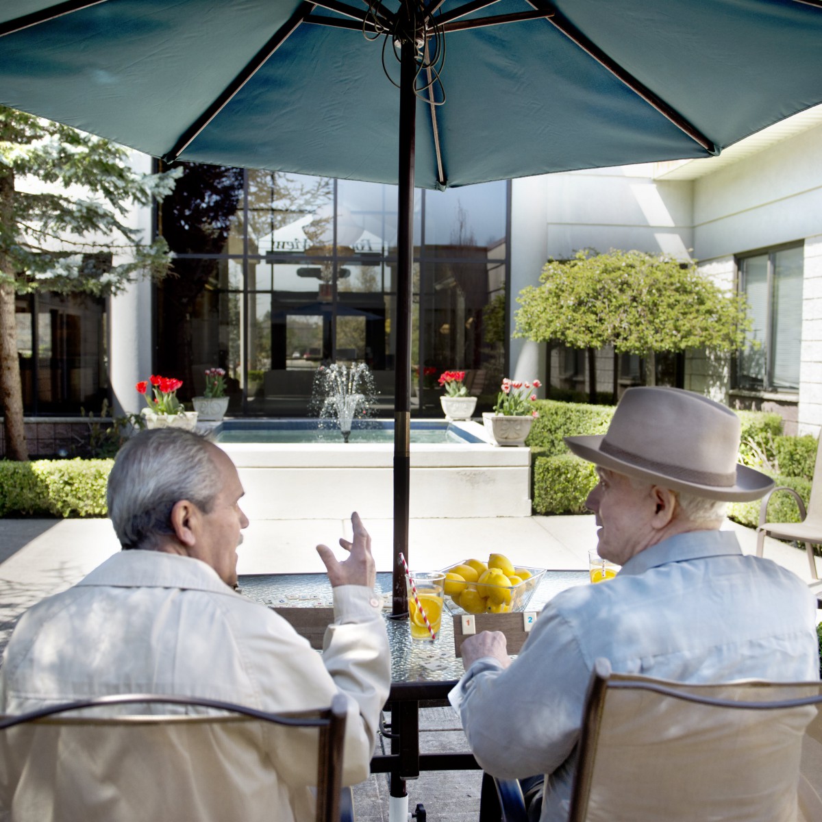 Two elderly individuals sitting under an umbrella, engaged in conversation on a patio.