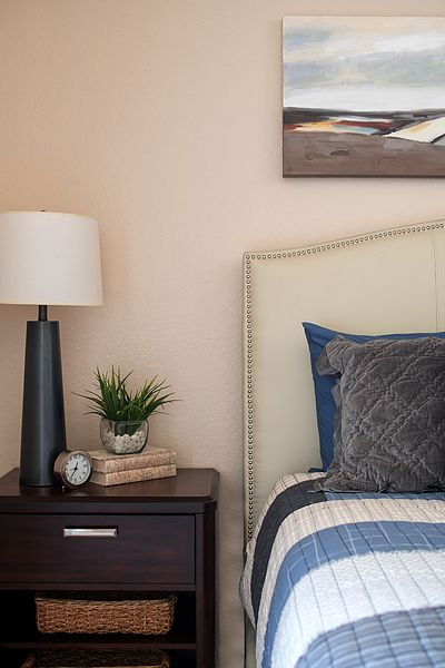 A cozy bedroom setup featuring a nightstand with a lamp, plant, books, and clock next to a bed with decorative pillows.