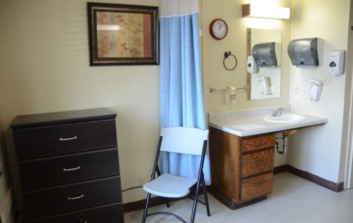 A small room featuring a folding chair, dresser, sink area with a mirror, and a curtain.