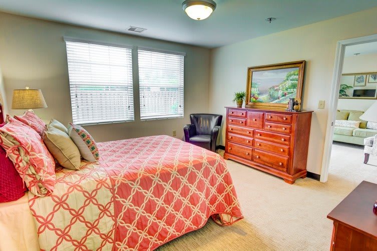 A cozy bedroom featuring a bed with a patterned comforter, a wooden dresser with a painting on top, and a chair in the corner.