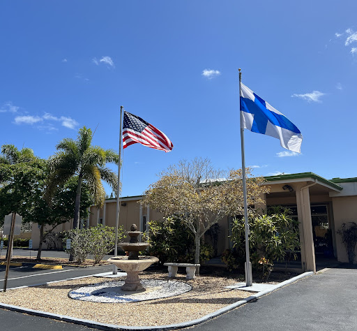 A view of a building with an American flag and a Finnish flag flying in front, accompanied by palm trees and a fountain.