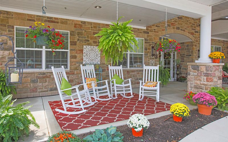A cozy porch with white rocking chairs, a red patterned rug, and vibrant hanging and potted plants.