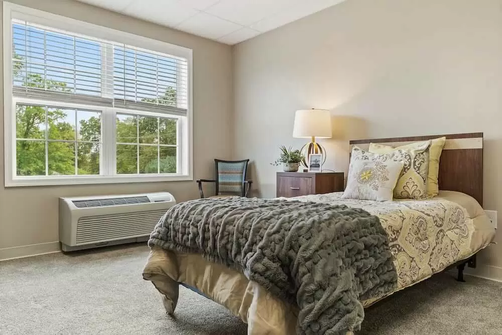 A cozy bedroom with a large window, featuring a neatly made bed, bedside table with lamp, and elegant decor.