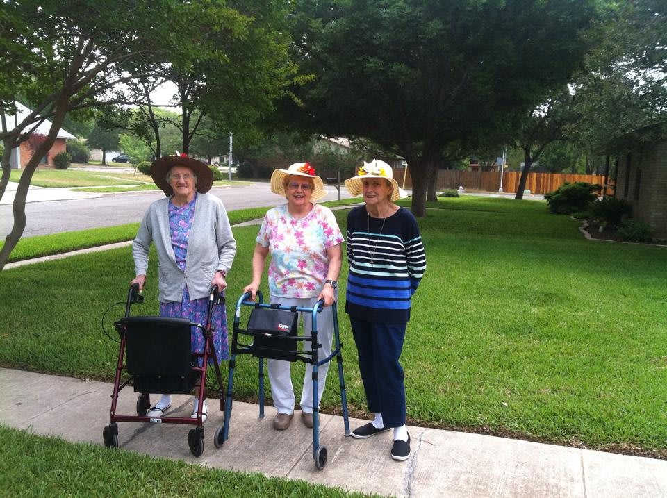 Three elderly women, wearing hats, enjoying a sunny walk outdoors with walkers.