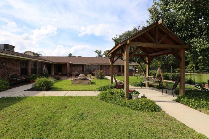 A peaceful garden setting with a wooden gazebo and lush greenery in front of a single-story house.