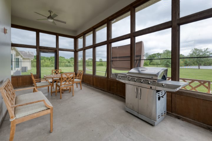 A modern screened porch with a stainless steel grill and wooden furniture set, overlooking a lush green landscape.