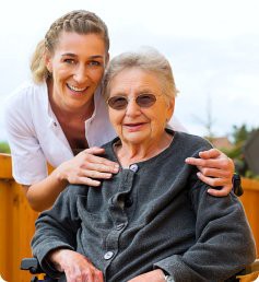 A caregiver smiling alongside an elderly person in a wheelchair outdoors.