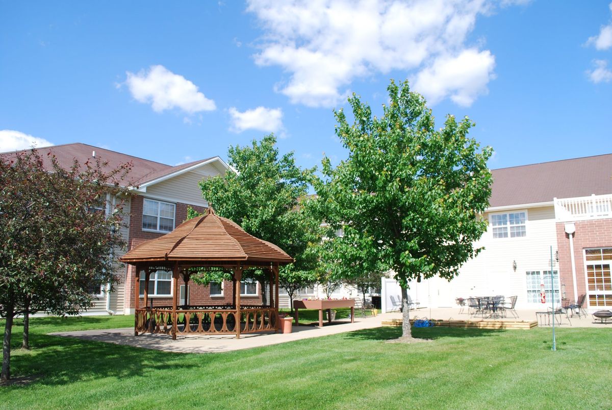 A scenic view of a wooden gazebo on a lush green lawn, surrounded by trees and overlooked by residential buildings under a clear blue sky.
