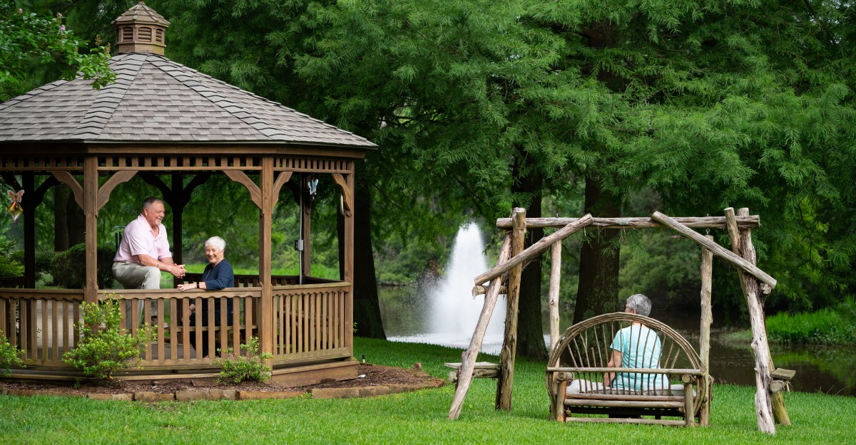 An outdoor scene featuring a gazebo and a swing with two people, set against a backdrop of trees and a fountain.
