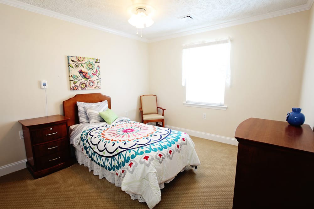 A cozy bedroom with a single bed adorned with colorful bedding, featuring a wooden chair and dresser, a carpeted floor, and bright wall art.