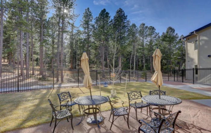 A scenic patio with wrought iron furniture and umbrellas set amidst a backdrop of lush trees and a clear blue sky.