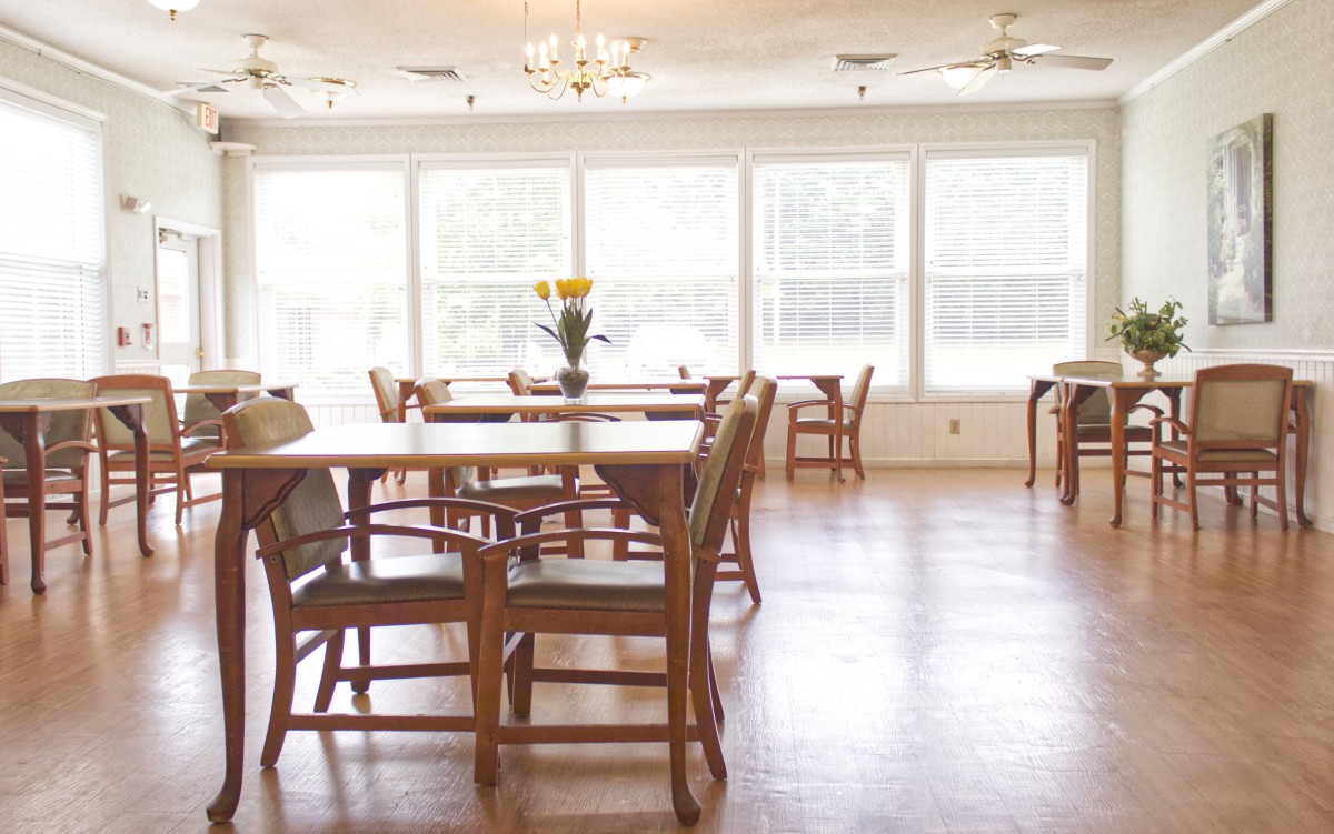 A bright and spacious dining room with wooden tables and chairs, featuring a vase with yellow flowers on one table and large windows letting in natural light.