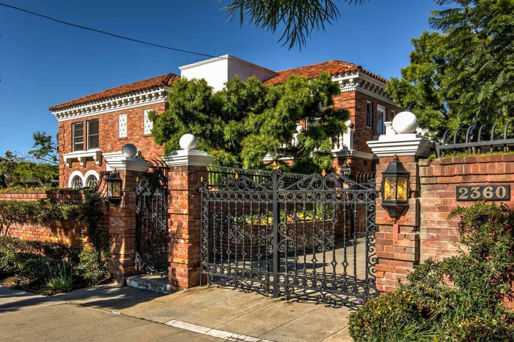 A large brick mansion with an ornate metal gate and surrounding greenery.