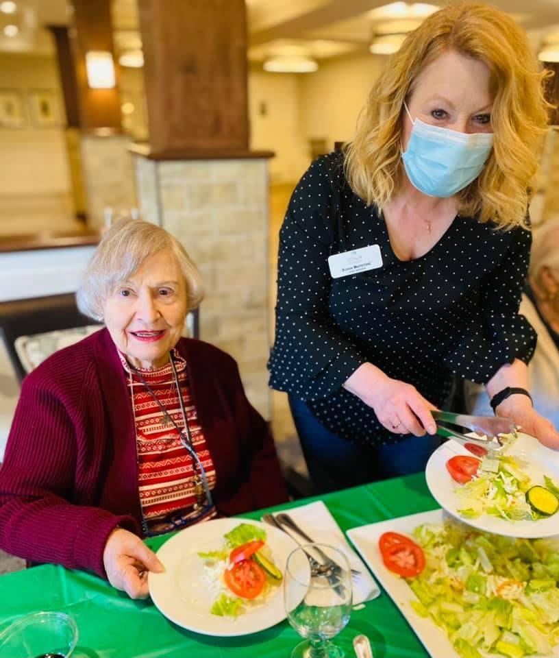 A caregiver serves a meal to an elderly woman at a dining area.