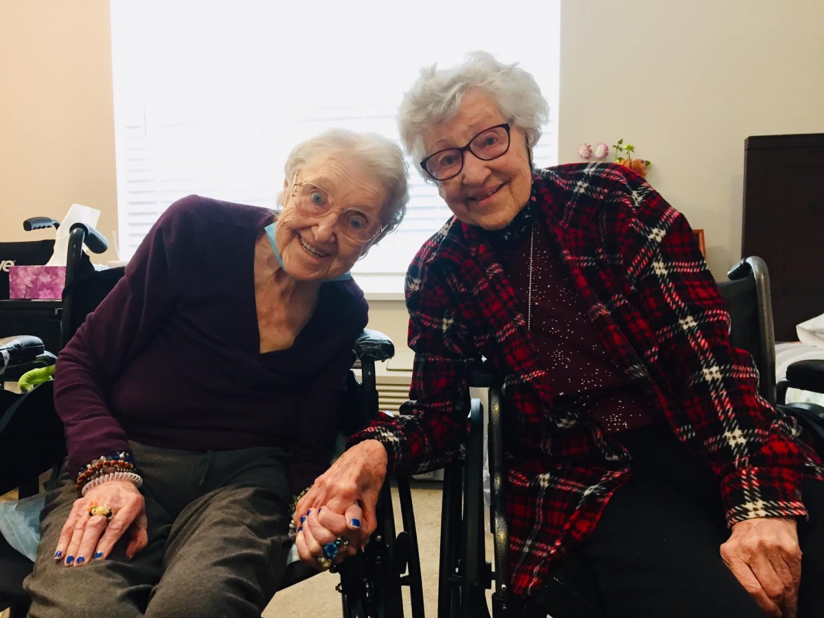 Two elderly women smiling and holding hands indoors, seated in wheelchairs.