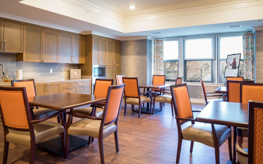 A cozy dining area with wooden tables and chairs, featuring a kitchen space and large windows.