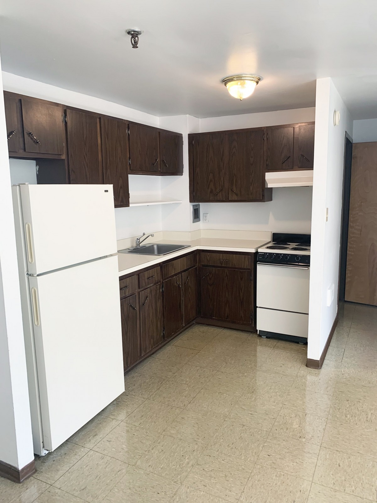 A simple apartment kitchen with dark wooden cabinets, a white fridge, and a stove.