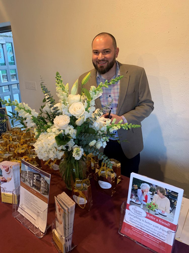 A man stands behind a table with a flower arrangement and books on display at an indoor event.