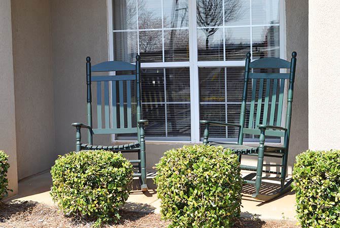 Two green rocking chairs on a patio in front of a window, surrounded by small bushes.