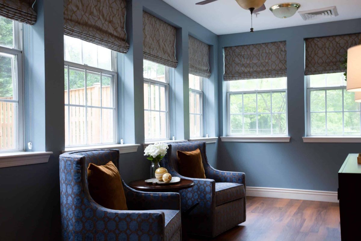 A cozy sitting area featuring two armchairs with accent pillows, surrounded by windows dressed in patterned curtains, complemented by elegant decor and wood flooring.