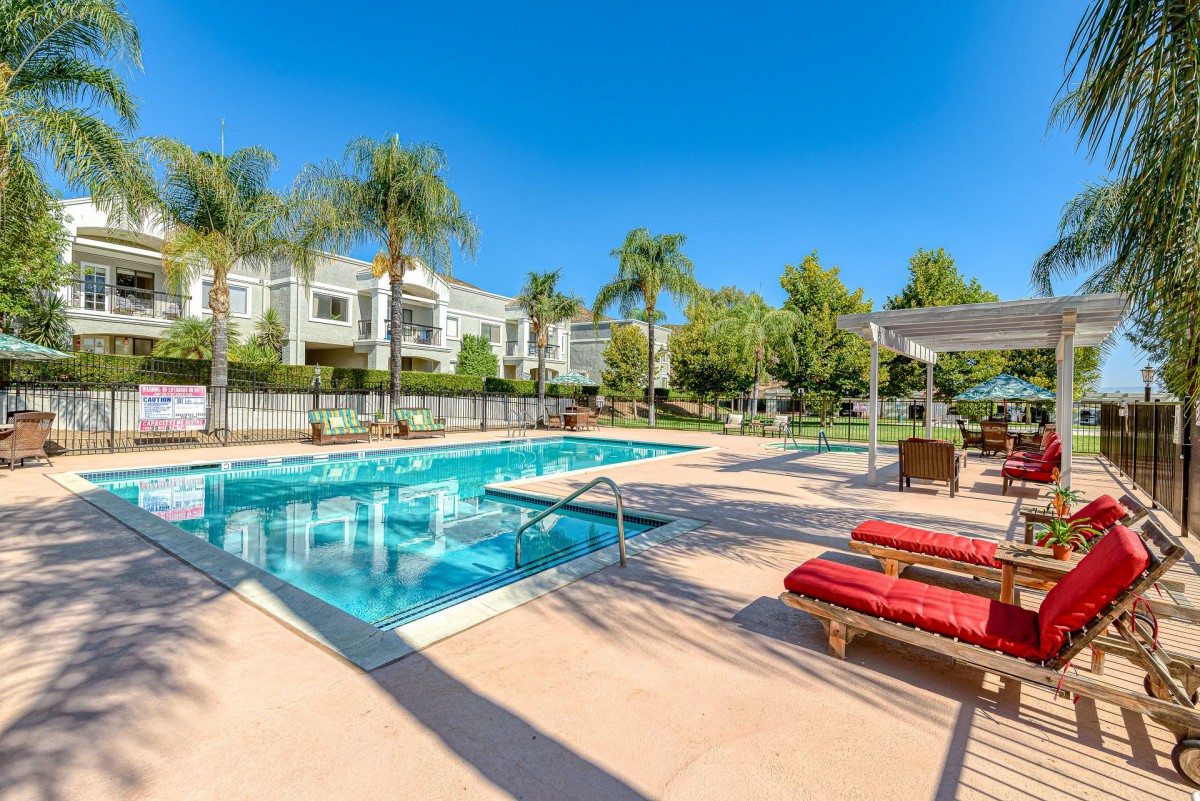 A serene residential swimming pool area surrounded by palm trees, featuring comfortable outdoor furniture under a bright blue sky.