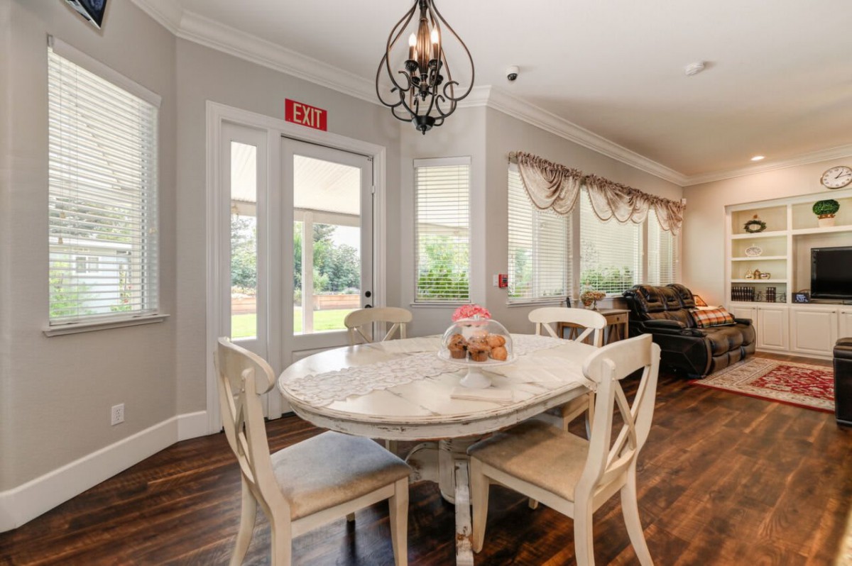 A cozy dining room with a round marble table and four chairs, featuring a decorative chandelier above and large windows providing natural light.