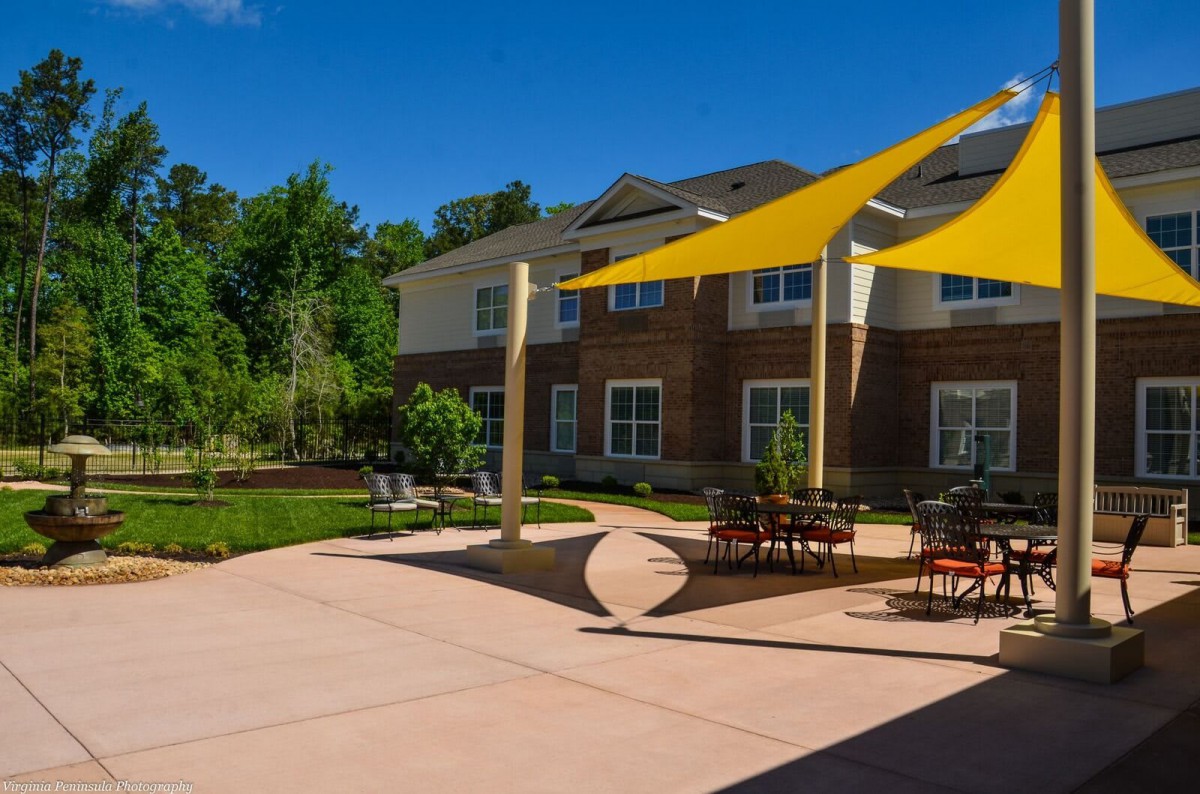A sunny patio with tables and chairs under yellow shade sails, adjacent to a building and surrounded by trees.