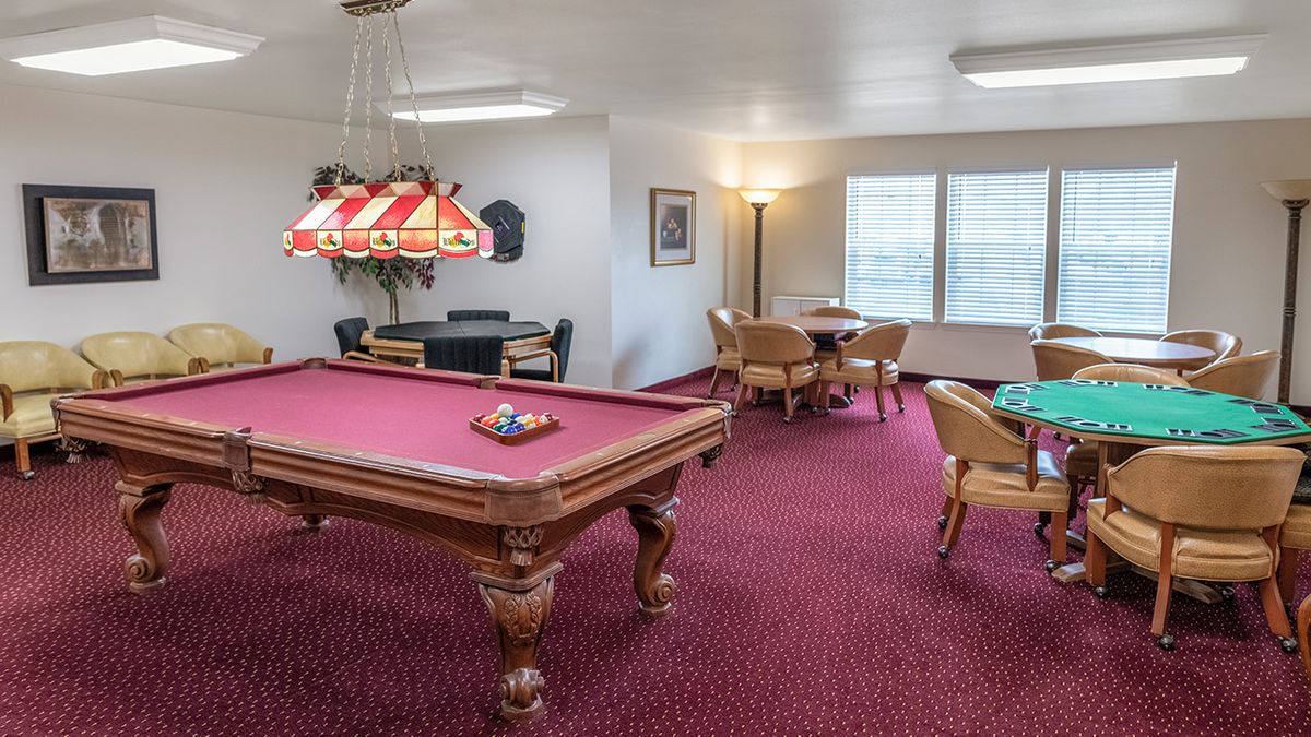 A game room featuring a billiards table with red felt, surrounded by seating and bright windows.
