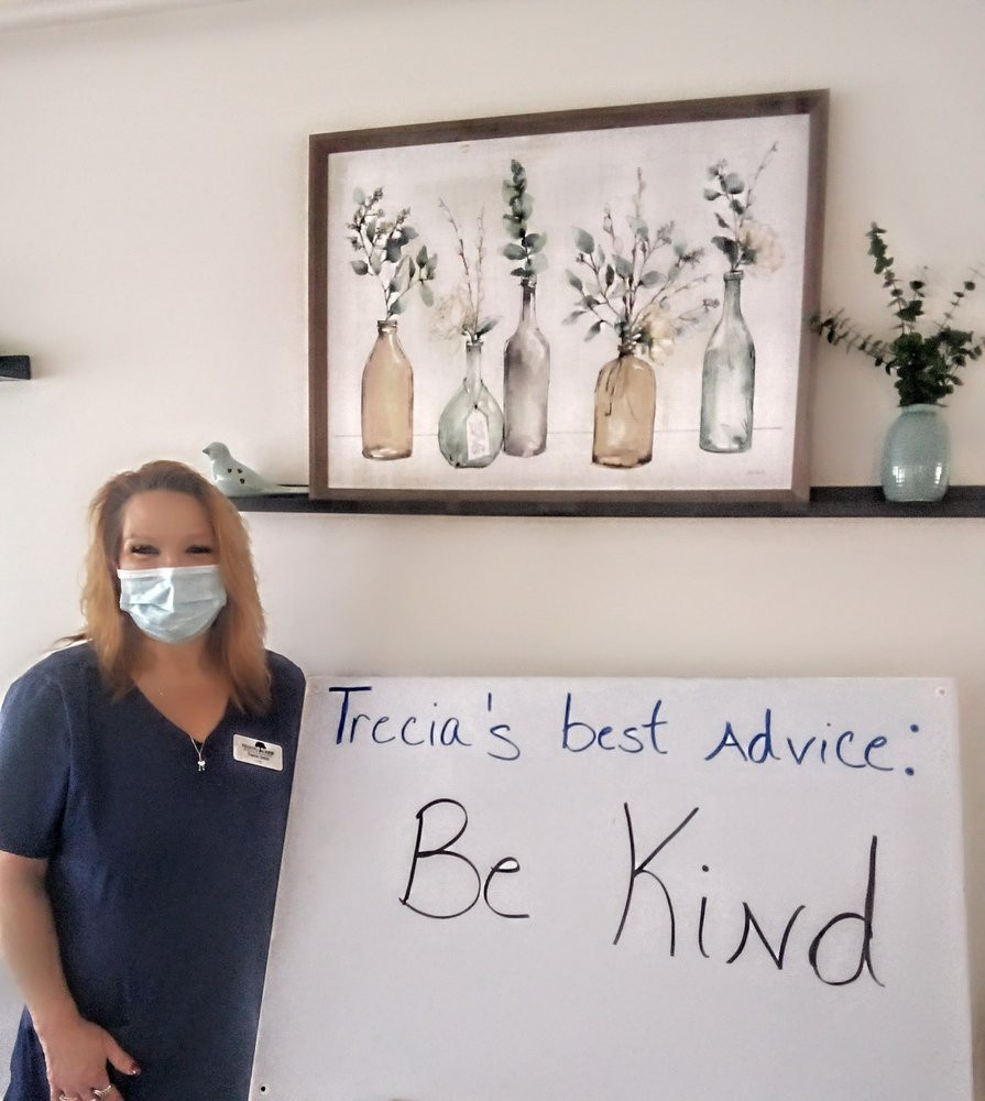 A healthcare worker wearing a mask stands beside a sign that says 'Trecia's best advice: Be Kind' with flower artwork in the background.