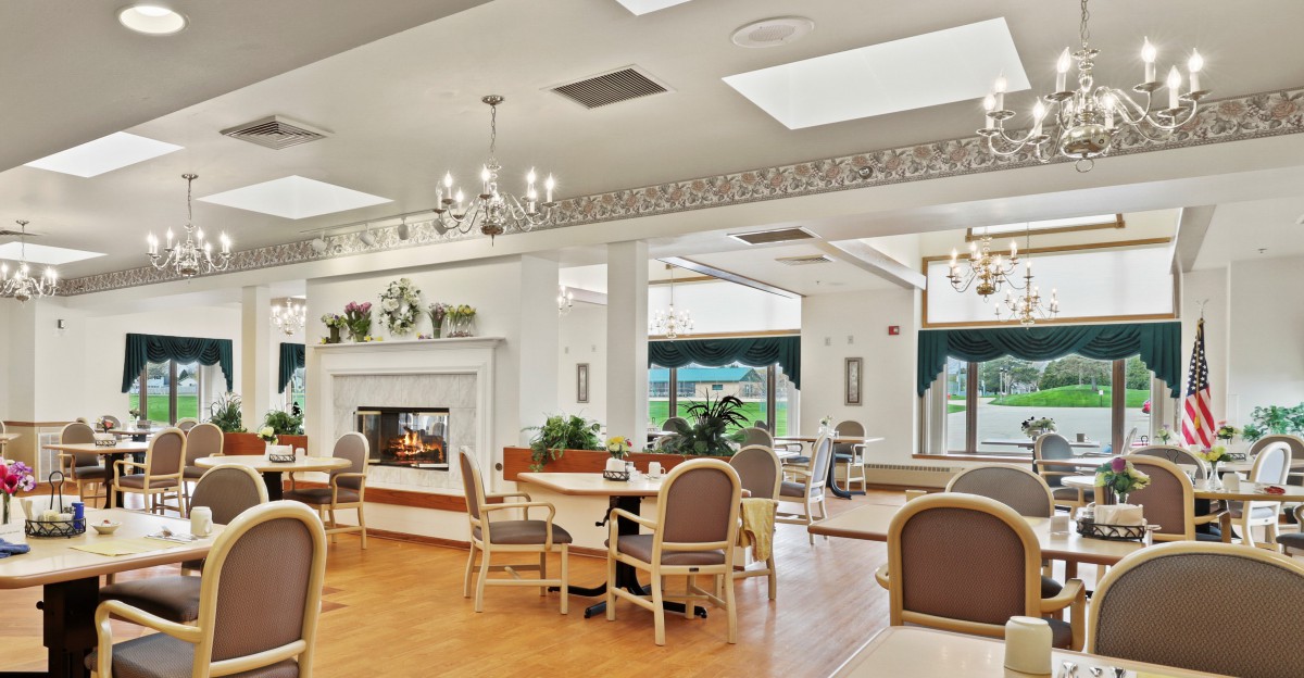 A cozy dining area with a fireplace, surrounded by tables and chairs, featuring natural light from large windows.