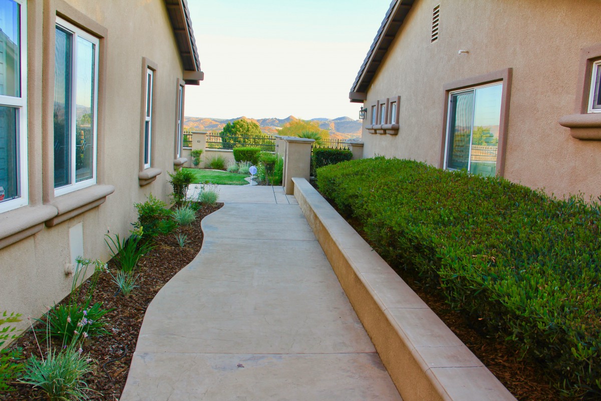 A walkway between two houses with shrubs and a view of the landscape beyond.