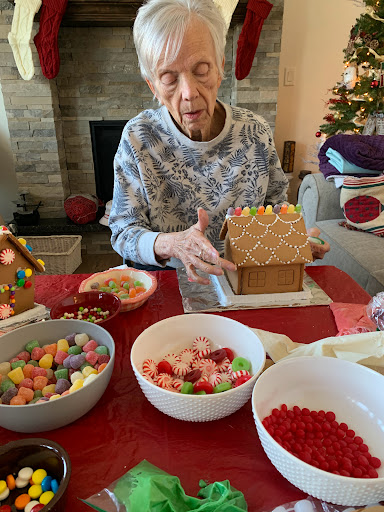 An elderly person joyfully decorating a gingerbread house surrounded by colorful candies during the festive season.