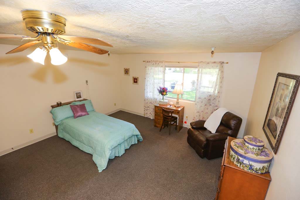 A cozy bedroom with a single bed, armchair, desk, and ceiling fan.