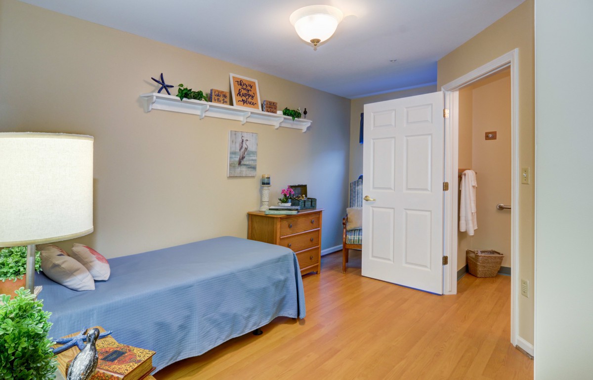 A cozy bedroom featuring a single bed with a blue cover, a dresser, and decorative items on a shelf. The room has wooden floors and an open door leading to another space.
