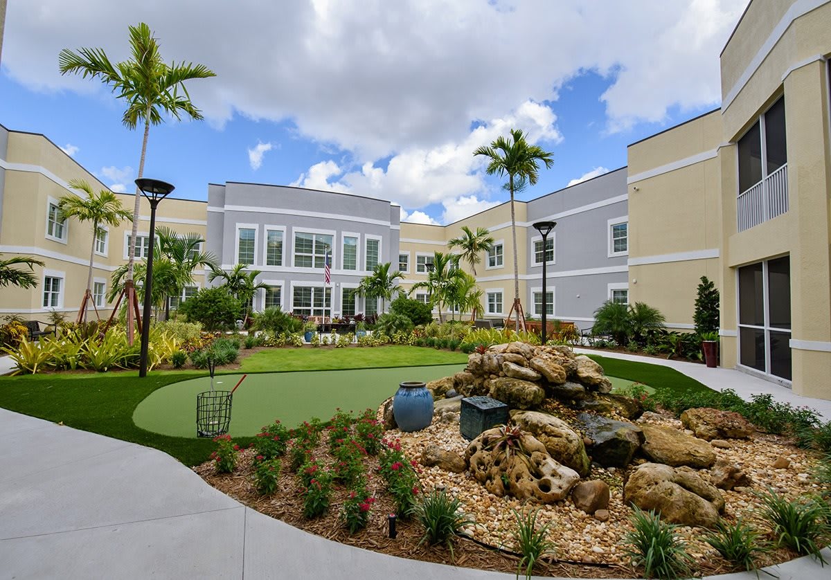 A modern building surrounded by a landscaped garden featuring palm trees and a stone decoration under a partly cloudy sky.
