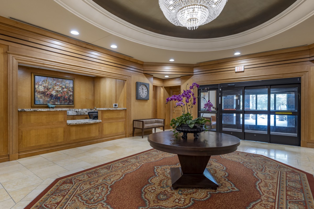 A luxurious hotel lobby featuring a grand chandelier, central table with a flower arrangement, and wooden paneling.