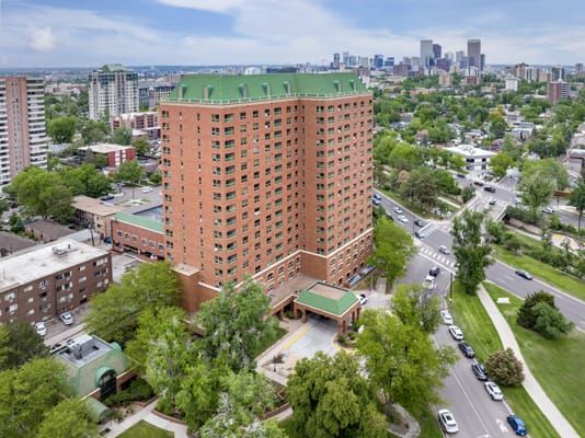 Aerial view of a tall brick apartment building surrounded by trees and roads, set against an urban cityscape.