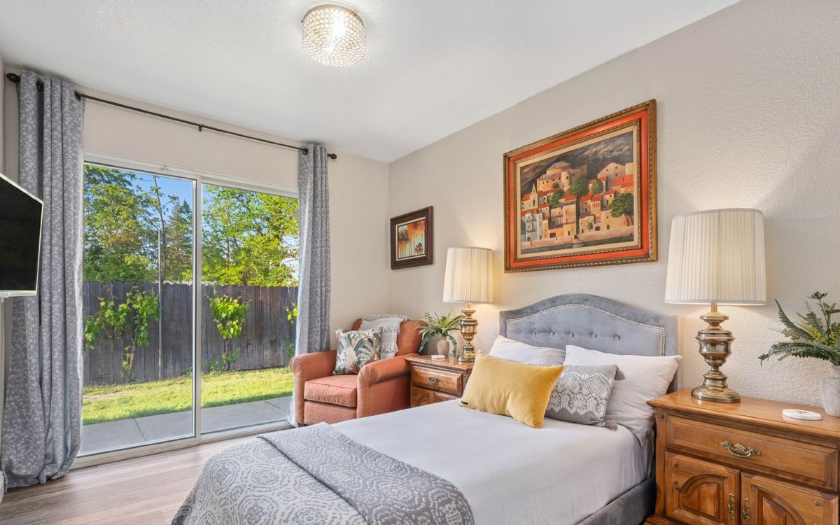 A cozy bedroom with a neatly made bed, an armchair by the window, and decorative paintings on the walls.