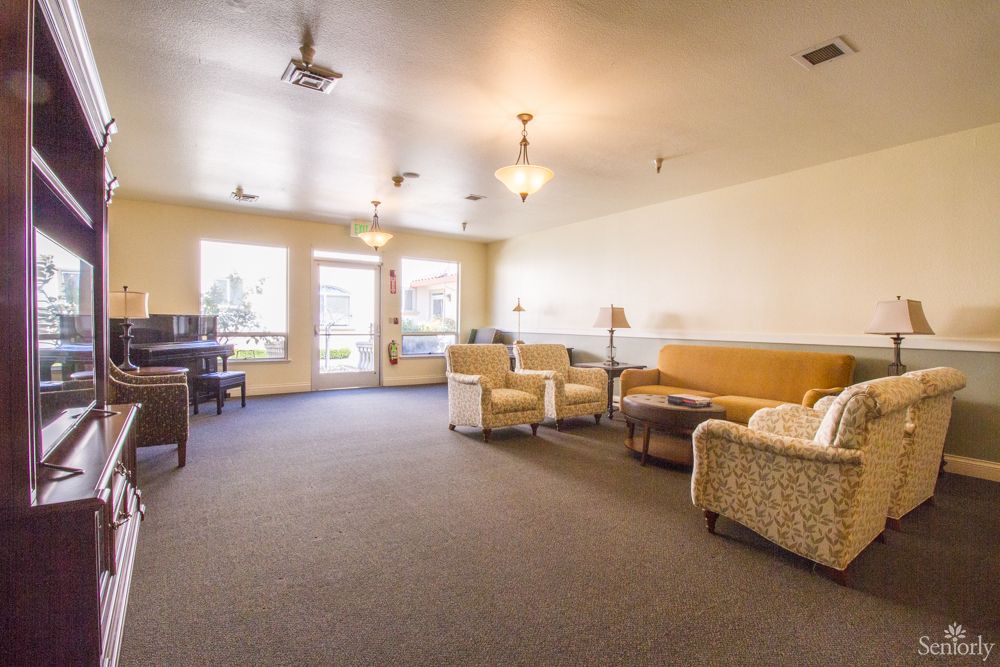 A spacious living room with comfortable seating, featuring sofas and armchairs, a piano, and large windows letting in natural light.