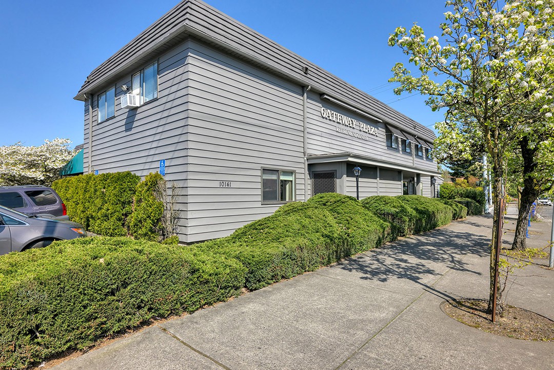 A two-story residential house with grey siding, surrounded by greenery and a sidewalk on a clear day.