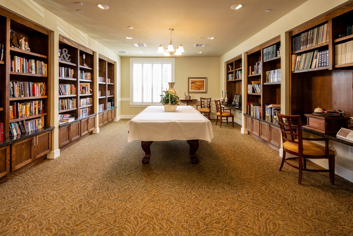 A cozy library room with bookshelves lining the walls and a large table in the center surrounded by chairs.