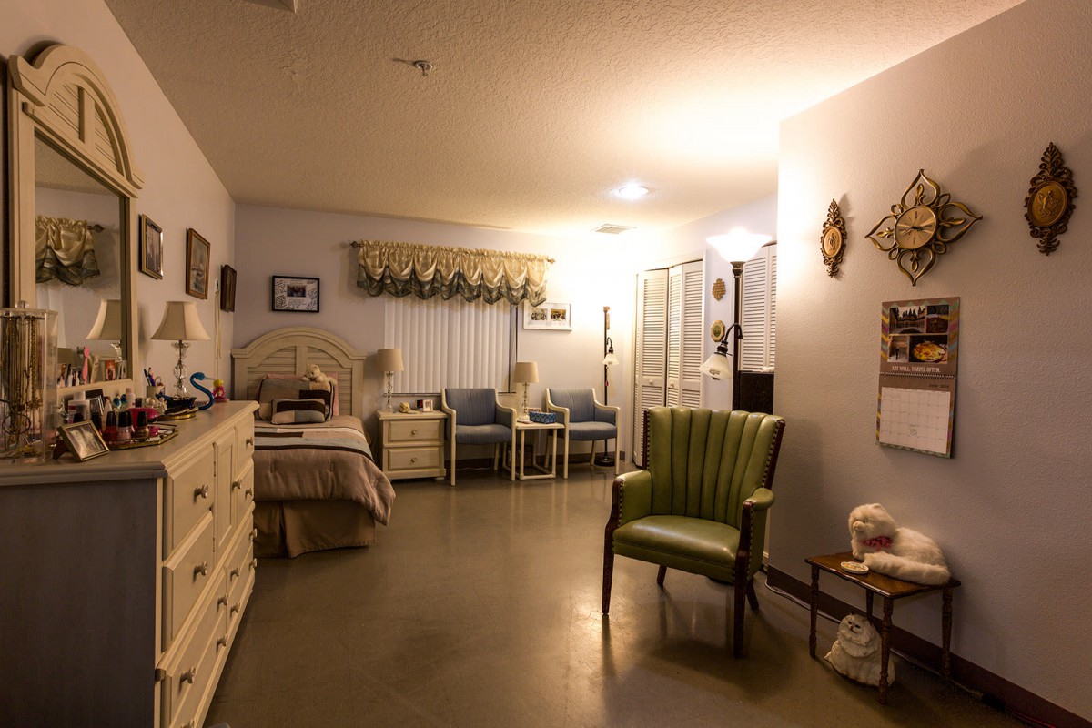 A cozy bedroom with a bed, dresser, and chairs, warmly lit by soft lighting.